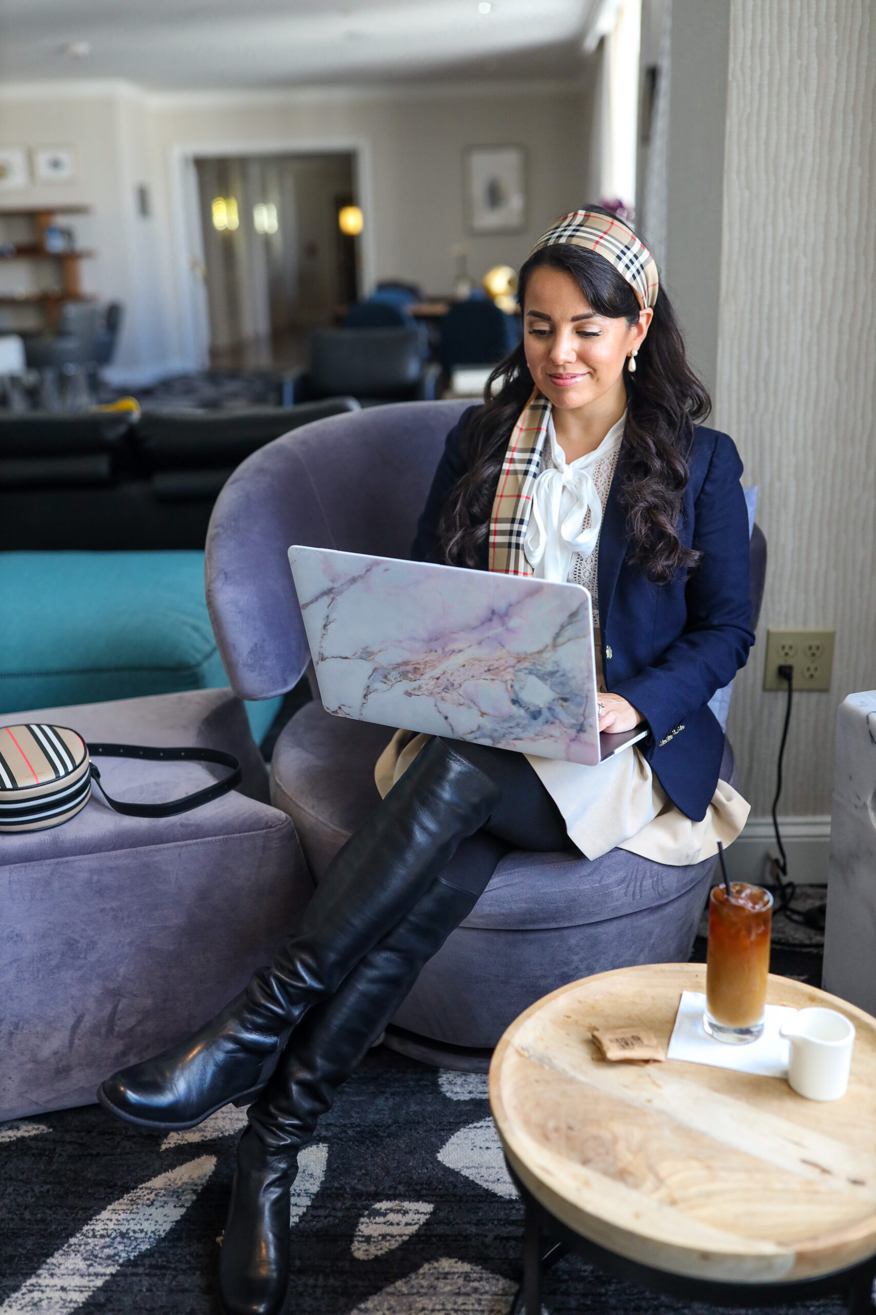 The Ritz-Carlton, San Francisco, Club Lounge, woman in navy blazer, beige pleated skirt and leather over-the-knee boots with matching Burberry plaid scarf and purse sits on purple chair with purple pastel laptop. Cold brew on table.