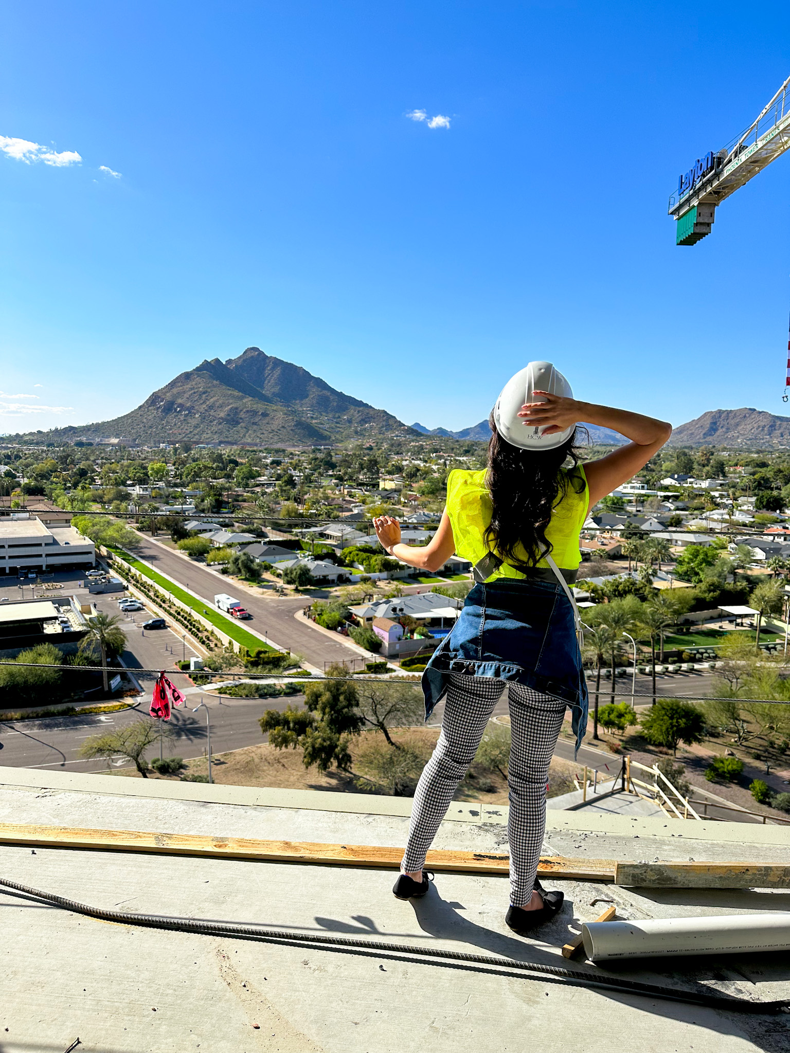 Woman stands with her back facing the camera in gingham pants, black shoes, green construction vest and white hard hat. She looks out to Camelback Mountain.