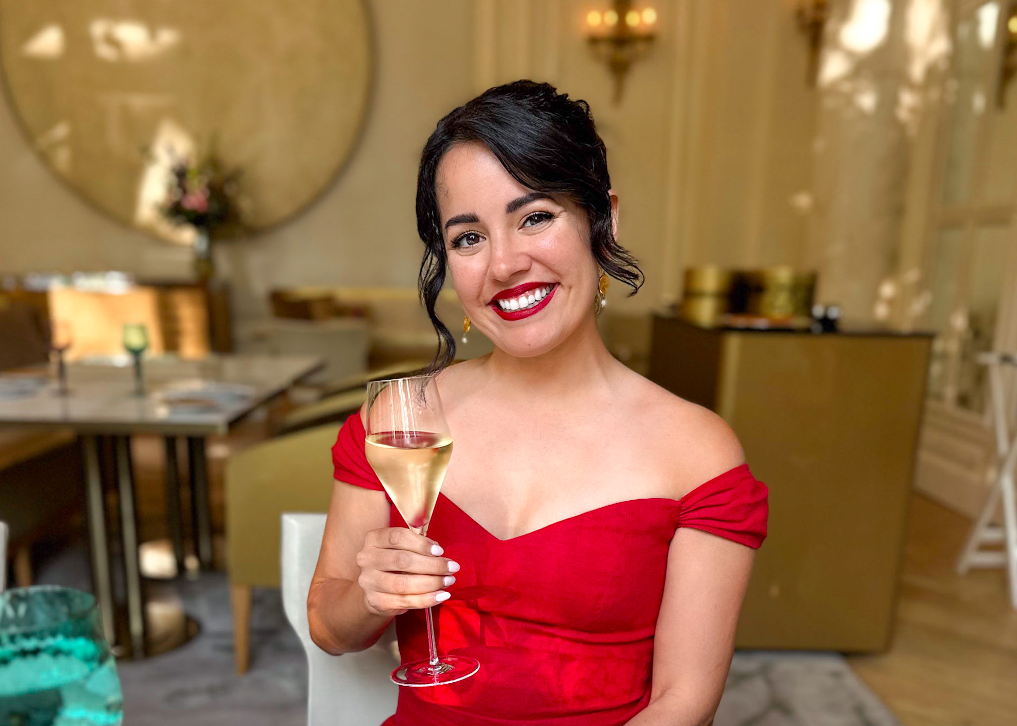 Natalie Moe, a dark-haired, olive skinned woman with an elegant up-do and a red off-shoulder dress, holds a glass of Ruinart Blanc de Blancs at the Mandarin-Oriental Ritz in Madrid, Spain.